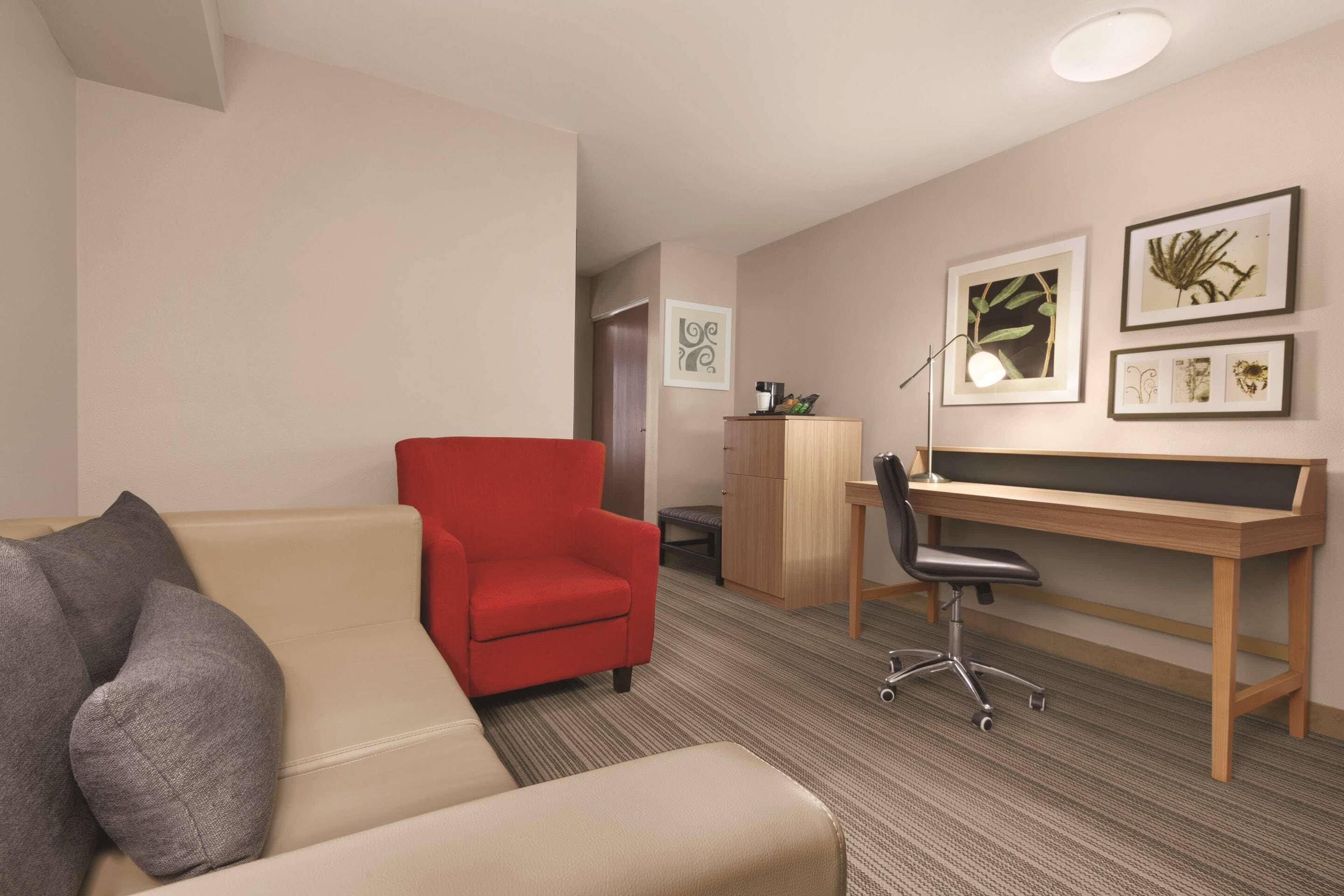 A hotel room with a beige sofa, a red armchair, a wooden desk, and a chair.