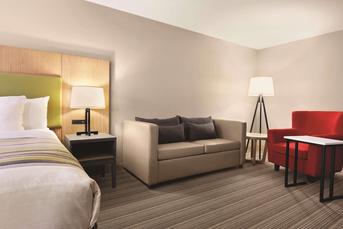 A hotel room with a bed, a sofa, a red armchair, a small table, and a floor lamp.