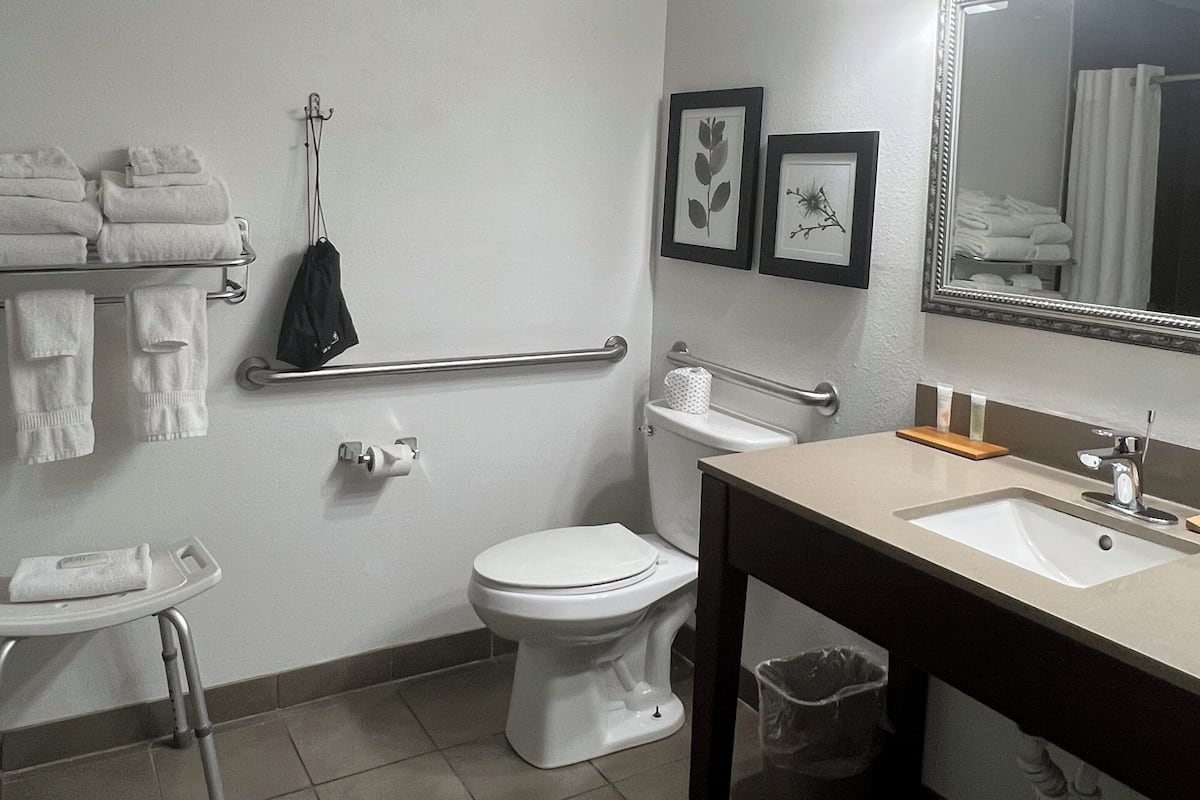 A standard hotel bathroom with a toilet, sink, and mirror. There are towels stacked on a shelf, a handrail, and a trash can.