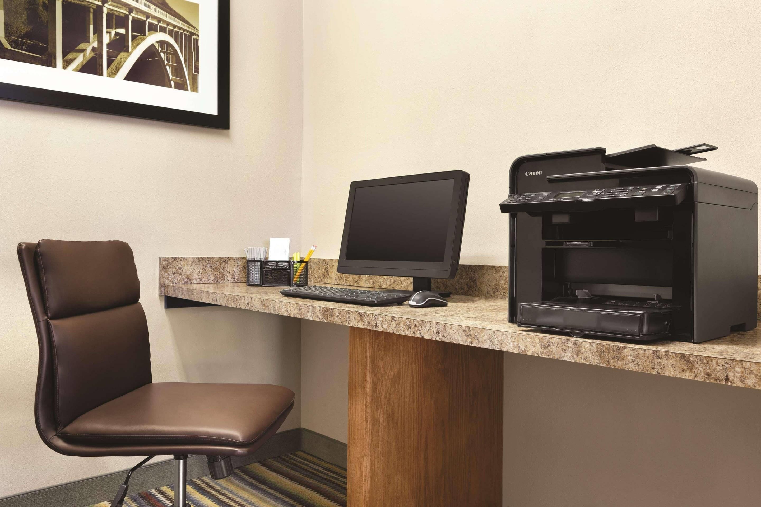 A hotel workspace with a desk, computer, printer, chair, and framed picture.