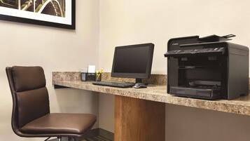 Un espacio de trabajo en un hotel con escritorio, computadora, impresora, silla e imagen enmarcada.