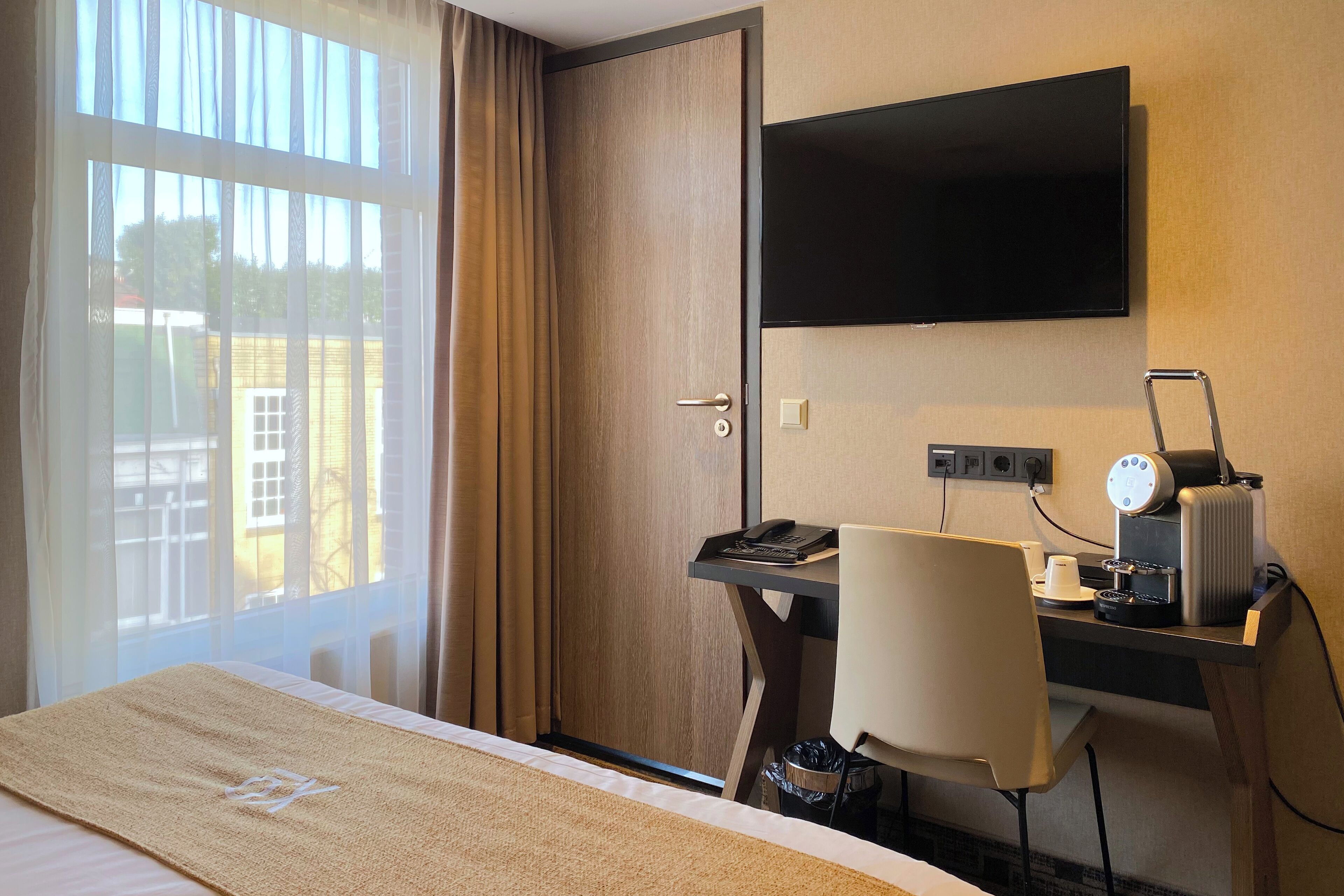 Una habitación de hotel con cama, escritorio y silla, televisor y ventana con cortinas.