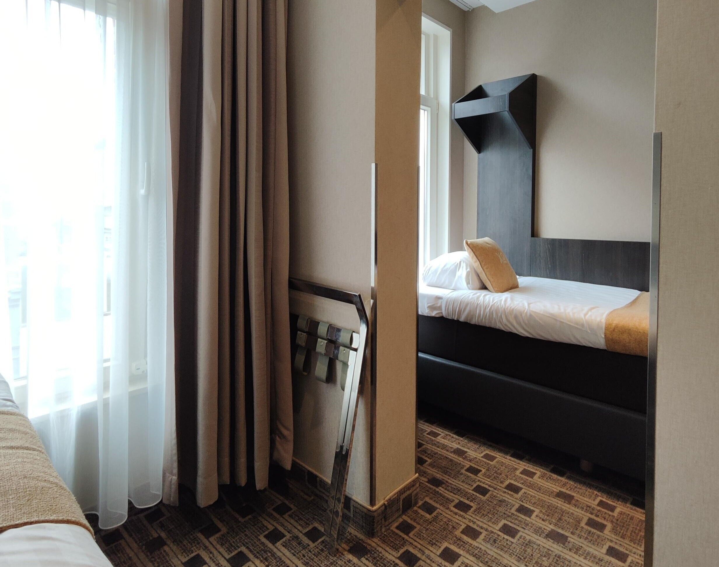 Een hotelkamer met een bed, een raam met gordijnen, een radiator en een wandplank.