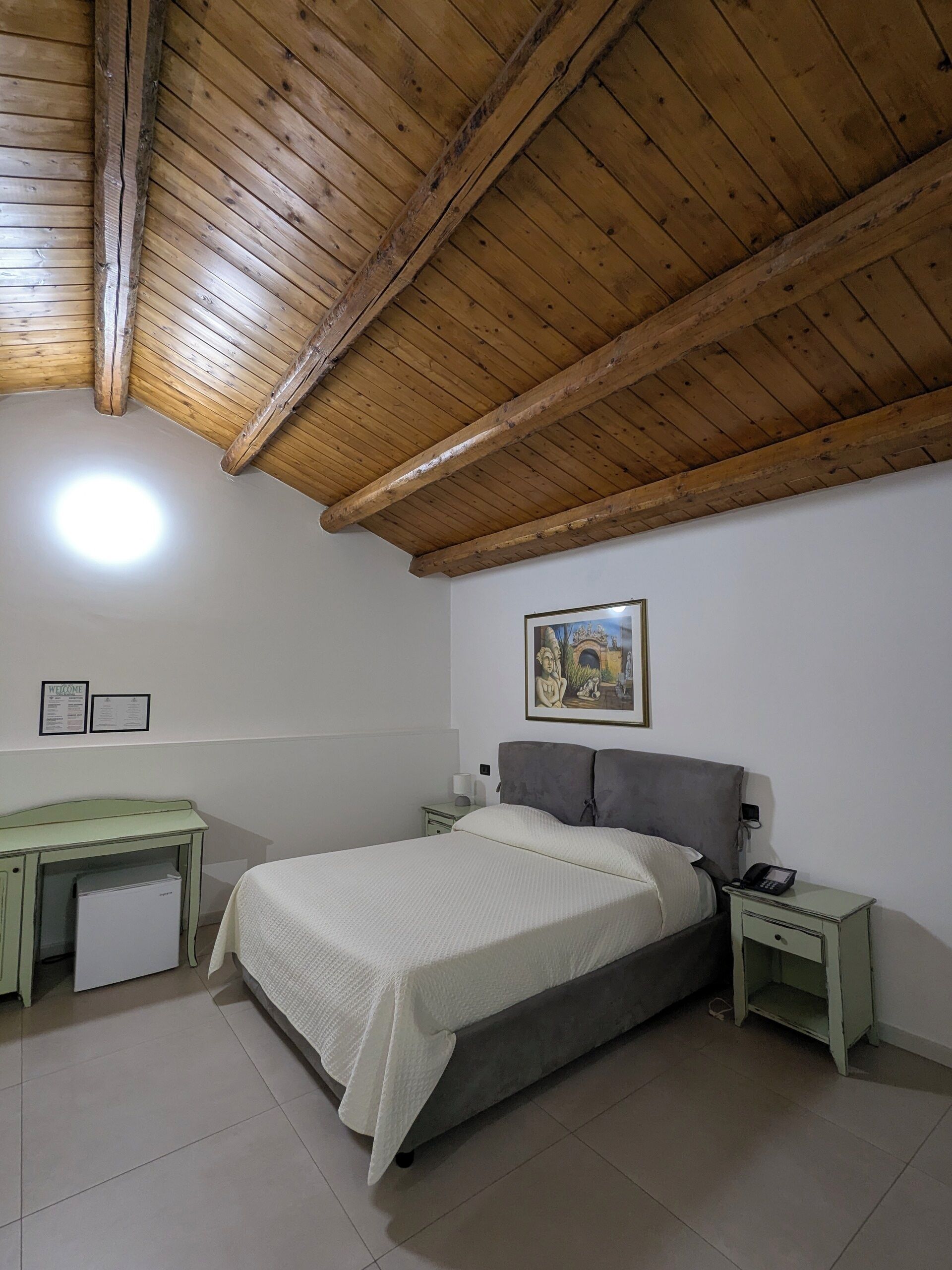 A bedroom with a bed, a desk, a nightstand, a painting, and a ceiling with wooden beams.