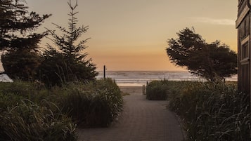 Un sentier pavé mène à une plage offrant une vue sur l’océan, bordé d’une végétation luxuriante et d’un bâtiment situé sur la droite.