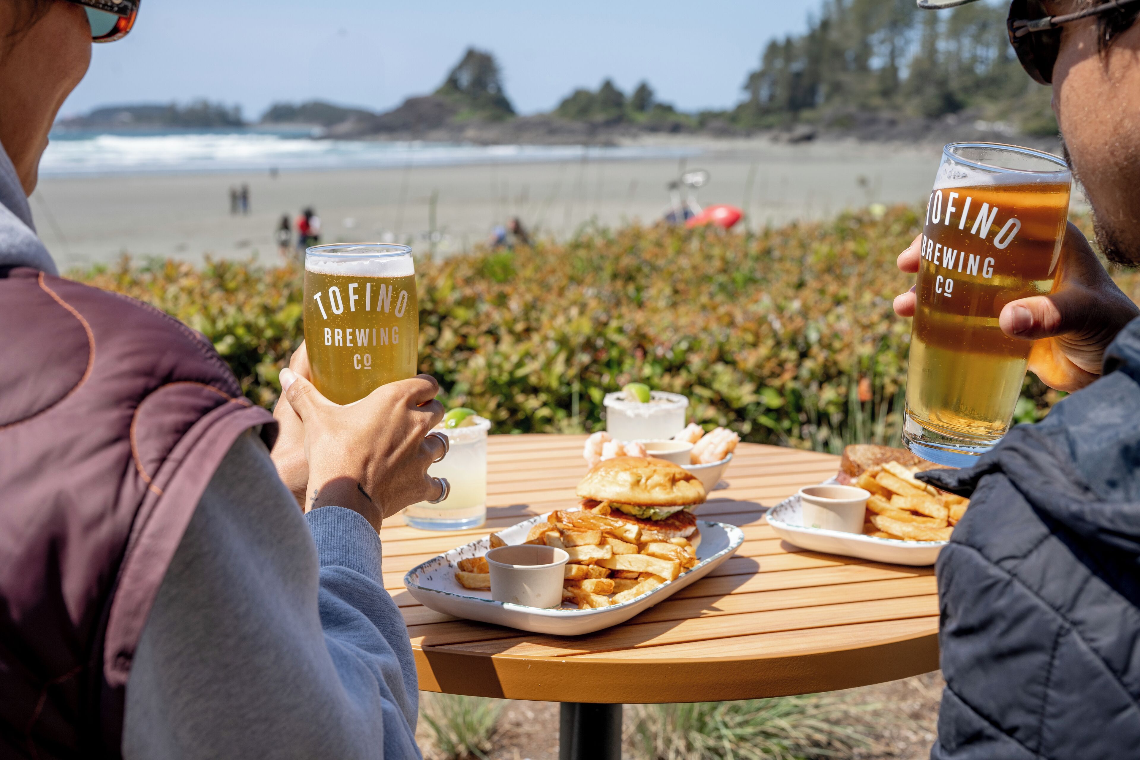 Dos personas sentadas en una mesa de madera con comida y bebidas, con vista a la playa.