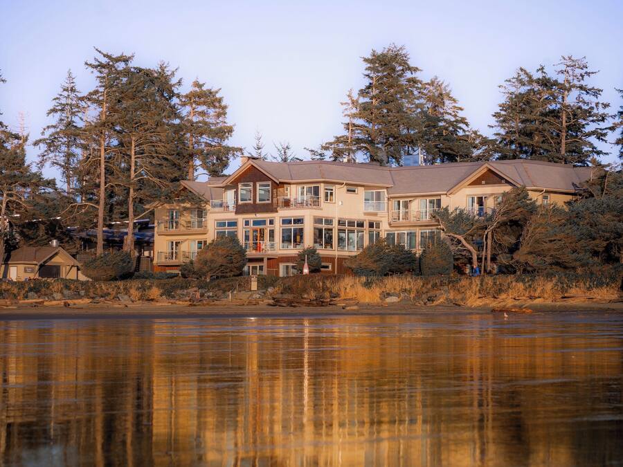 A large, multi-story house with a prominent balcony, situated by a lake with dense trees in the background.