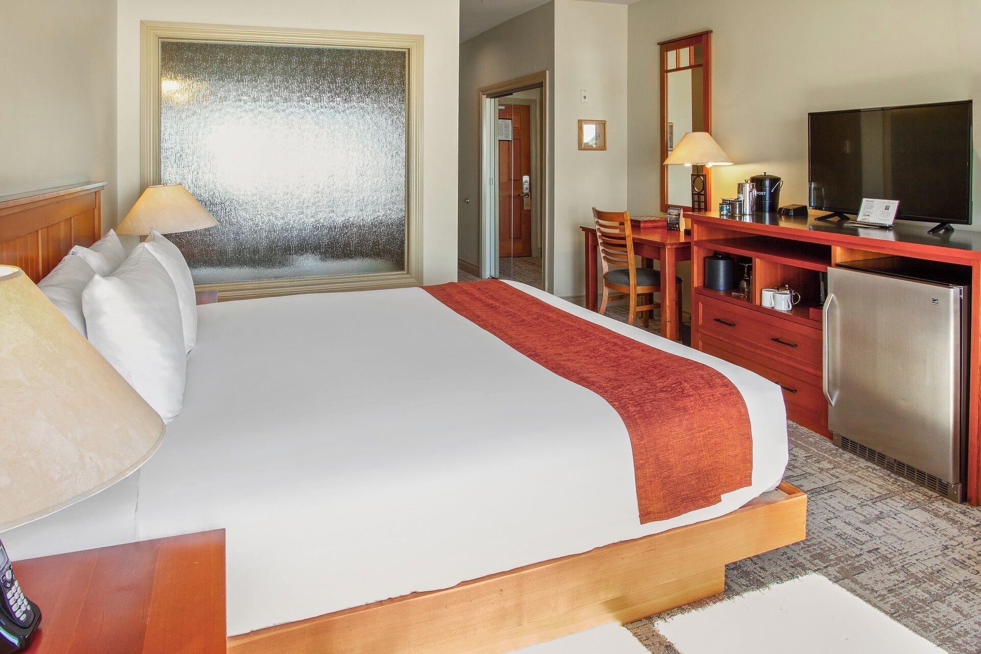 Ein Hotelzimmer mit einem großen Bett, einem Schreibtisch mit Fernseher, einem Kühlschrank und einem Fenster mit Milchglasverglasung.