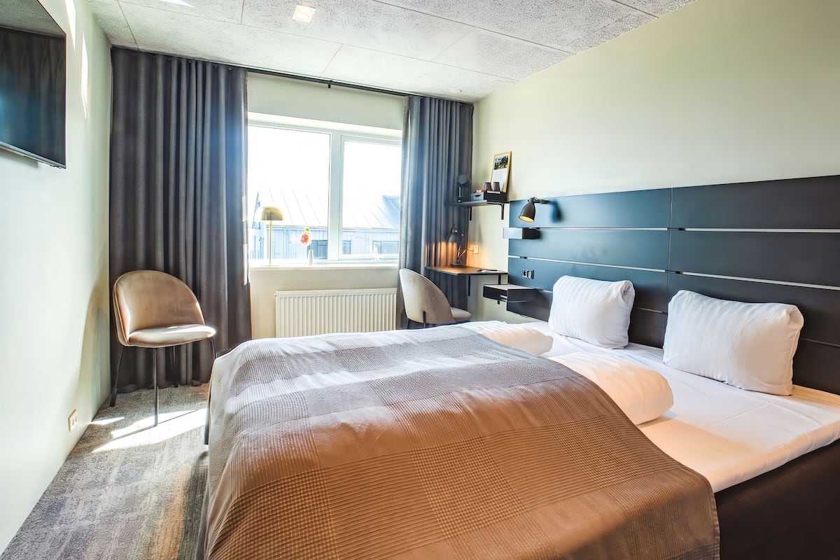 Une chambre d’hôtel avec un lit, une chaise, un bureau, une fenêtre avec des rideaux et un téléviseur fixé au mur.