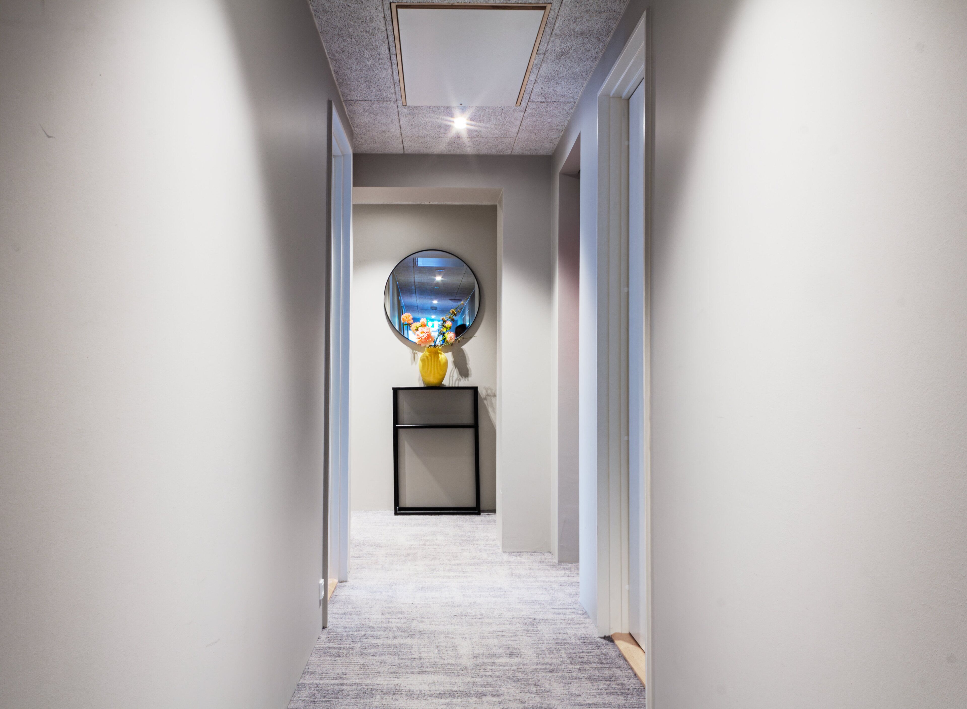 A hallway with a carpeted floor, a round mirror on a pedestal, and a framed picture on the wall.