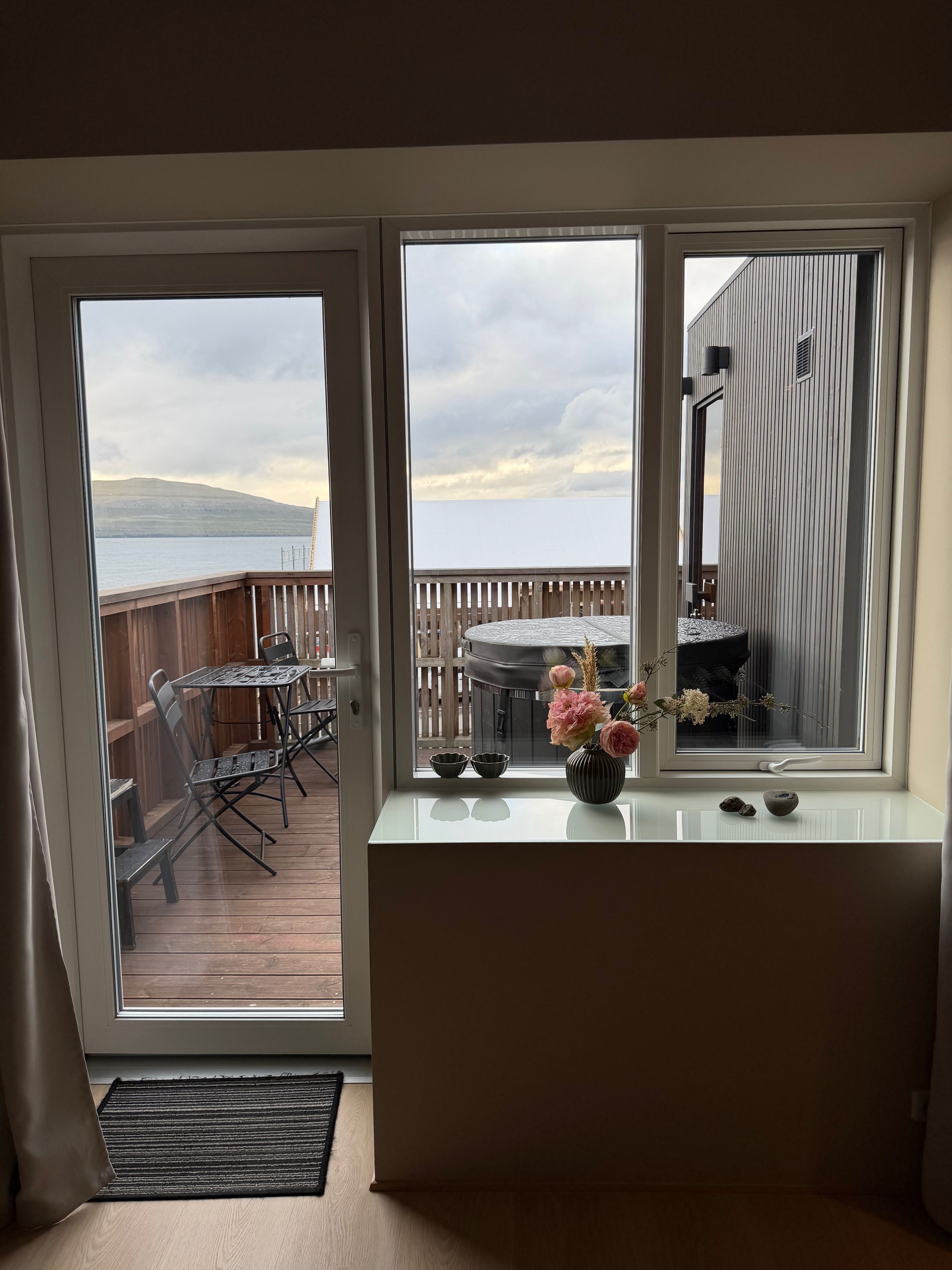 A room with a view of a wooden deck and a hot tub, a window with a vase of flowers, and a small table with chairs.
