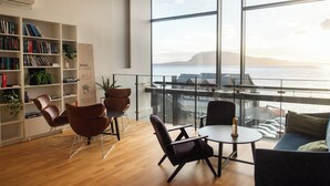A modern room with a large window overlooking the sea, a bookshelf, and seating area. - Hotel Djurhuus (Tórshavn)