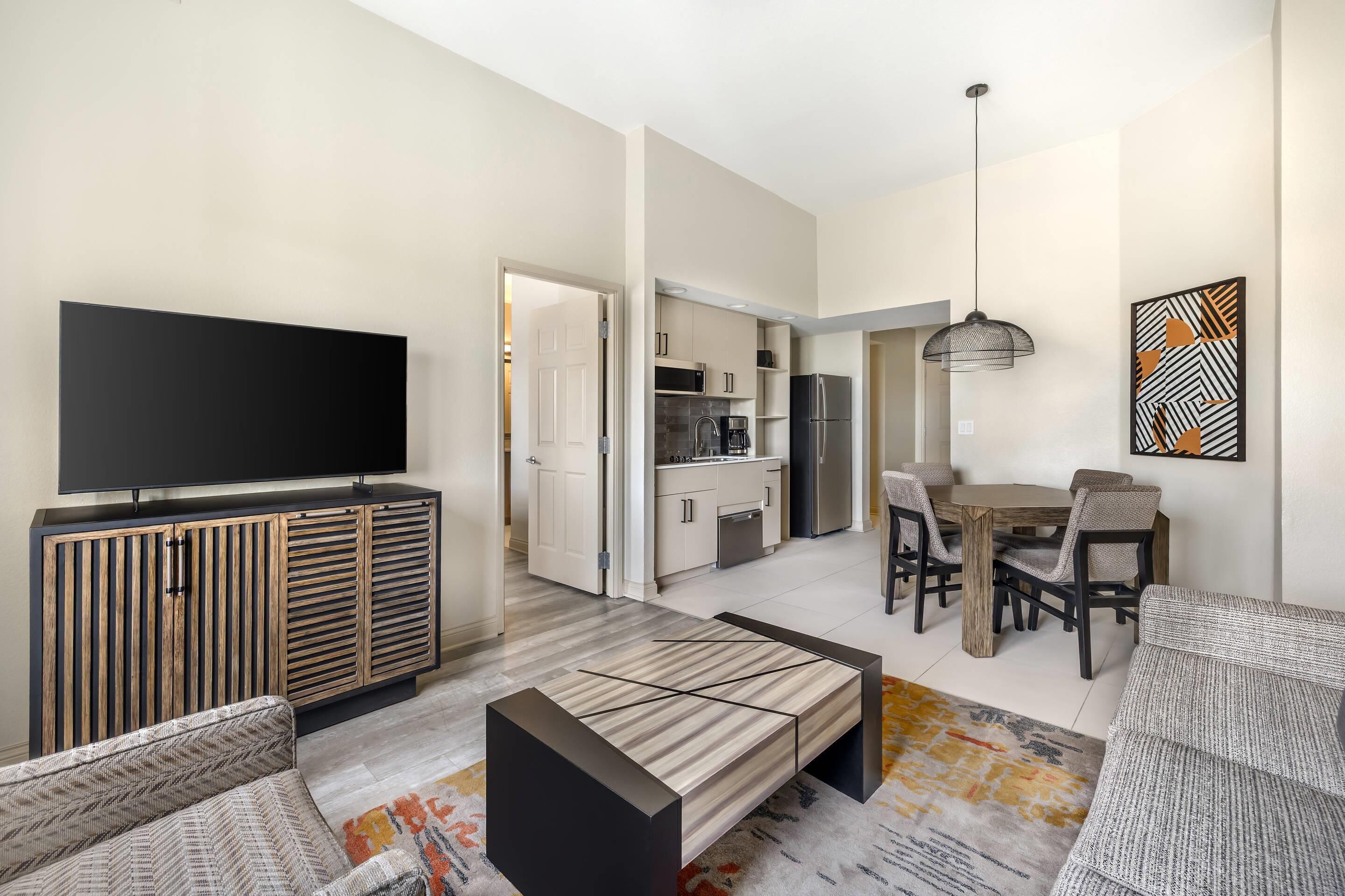 Een moderne woonkamer met een flatscreen televisie, een bank, een salontafel, een eethoek met een houten tafel en stoelen, en een keuken op de achtergrond.