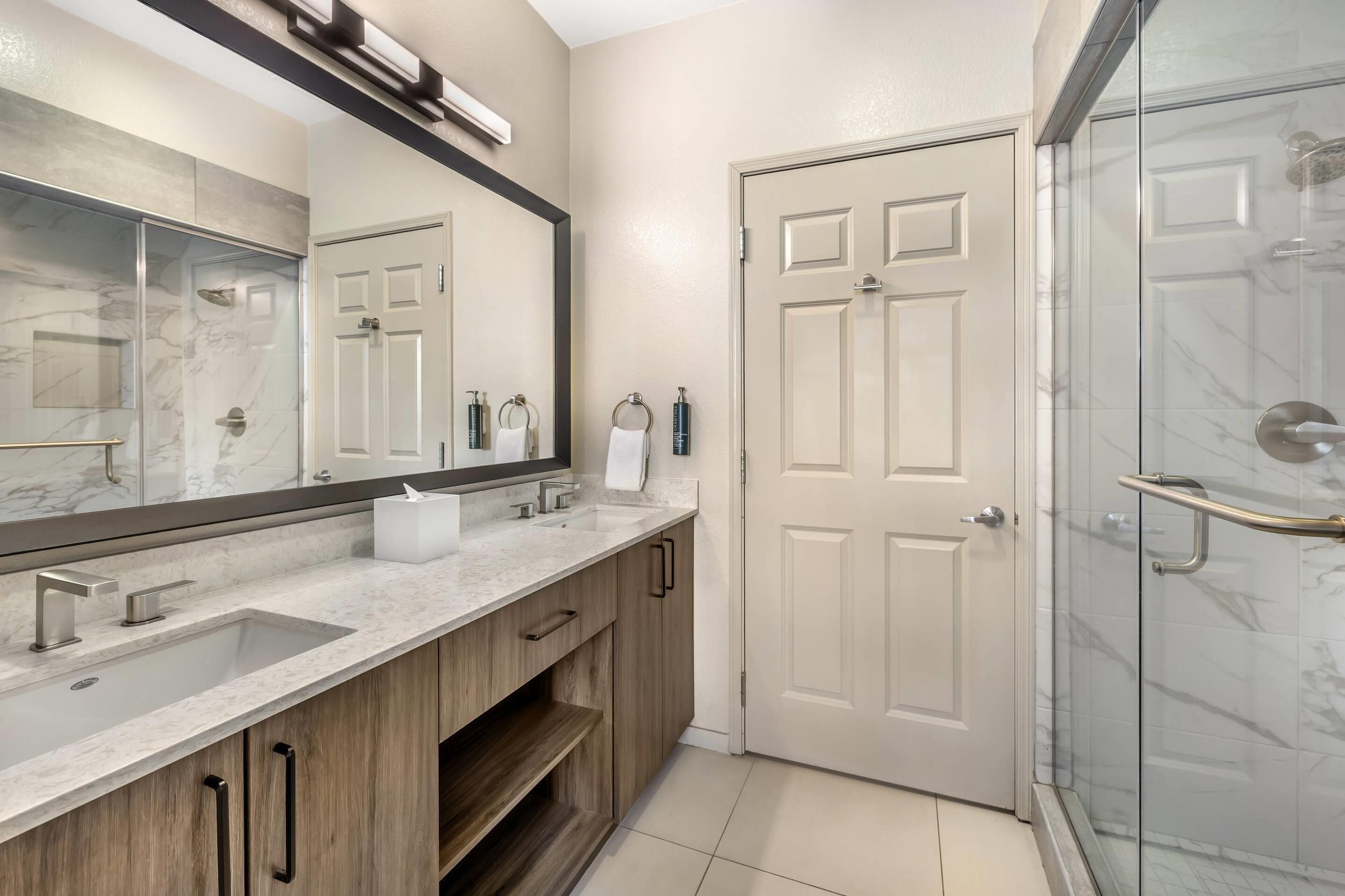 A bathroom with a marble sink, wooden cabinets, a large mirror, a glass shower enclosure, and a white door.