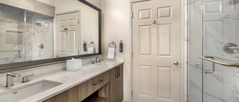 A bathroom with a marble sink, wooden cabinets, a large mirror, a glass shower enclosure, and a white door.