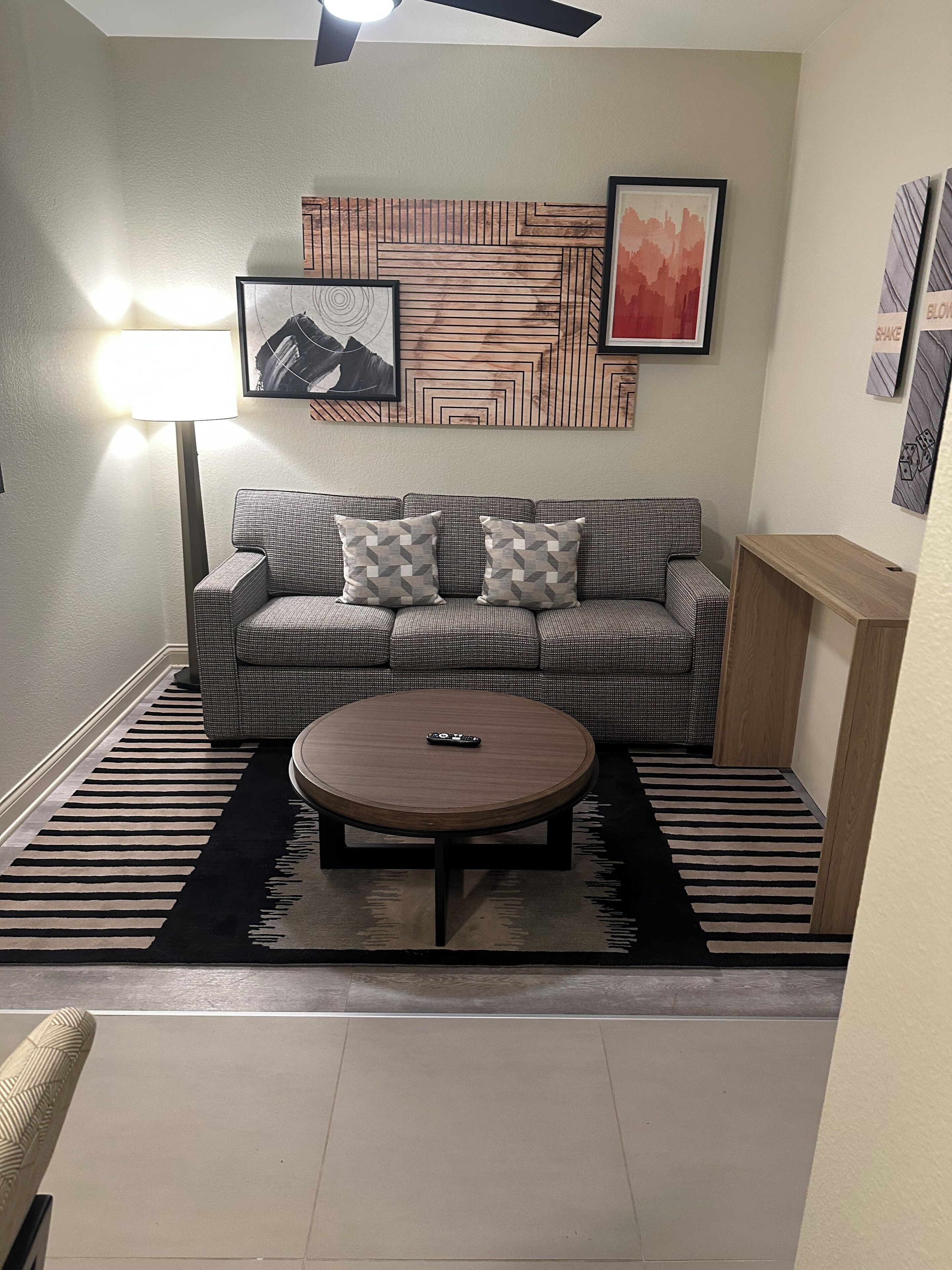 Un salon comprenant un canapé, deux coussins à motifs, une table basse ronde, un lampadaire et un mur orné de motifs géométriques et de tableaux encadrés.