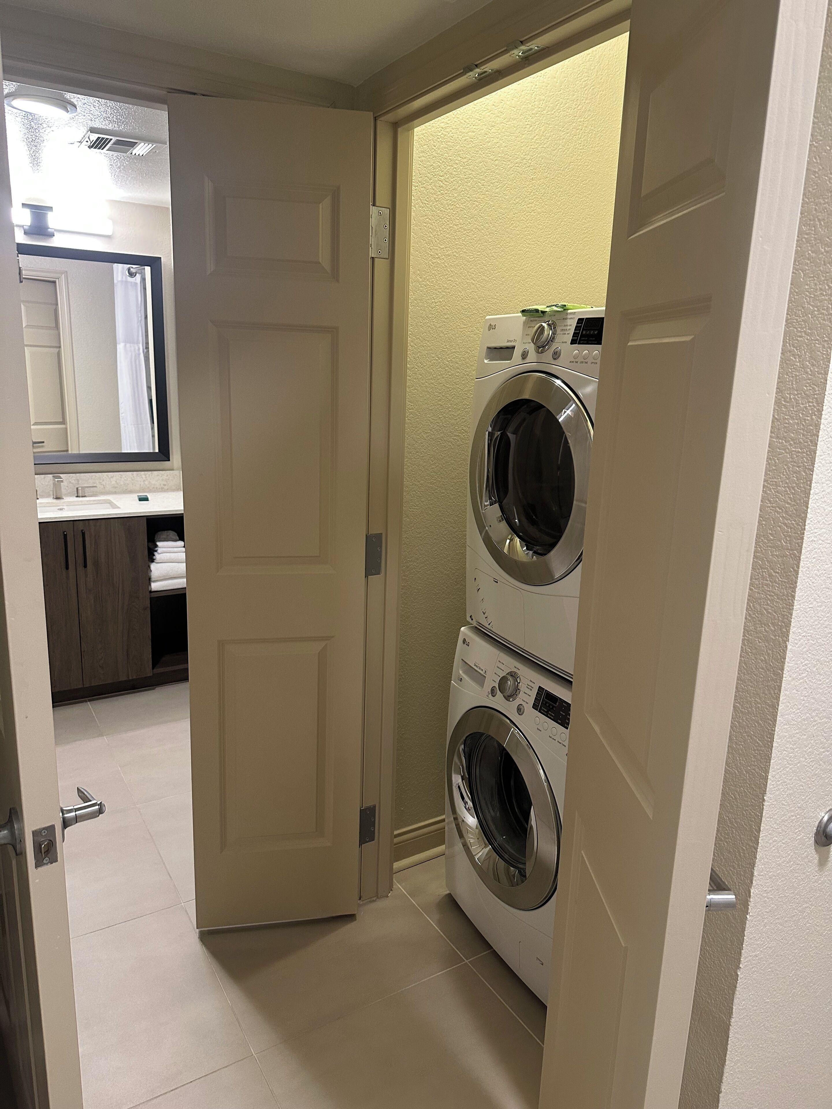 A laundry room with two stacked front-loading washers and dryers, a door leading to a bathroom, and a small countertop with a sink and mirror.