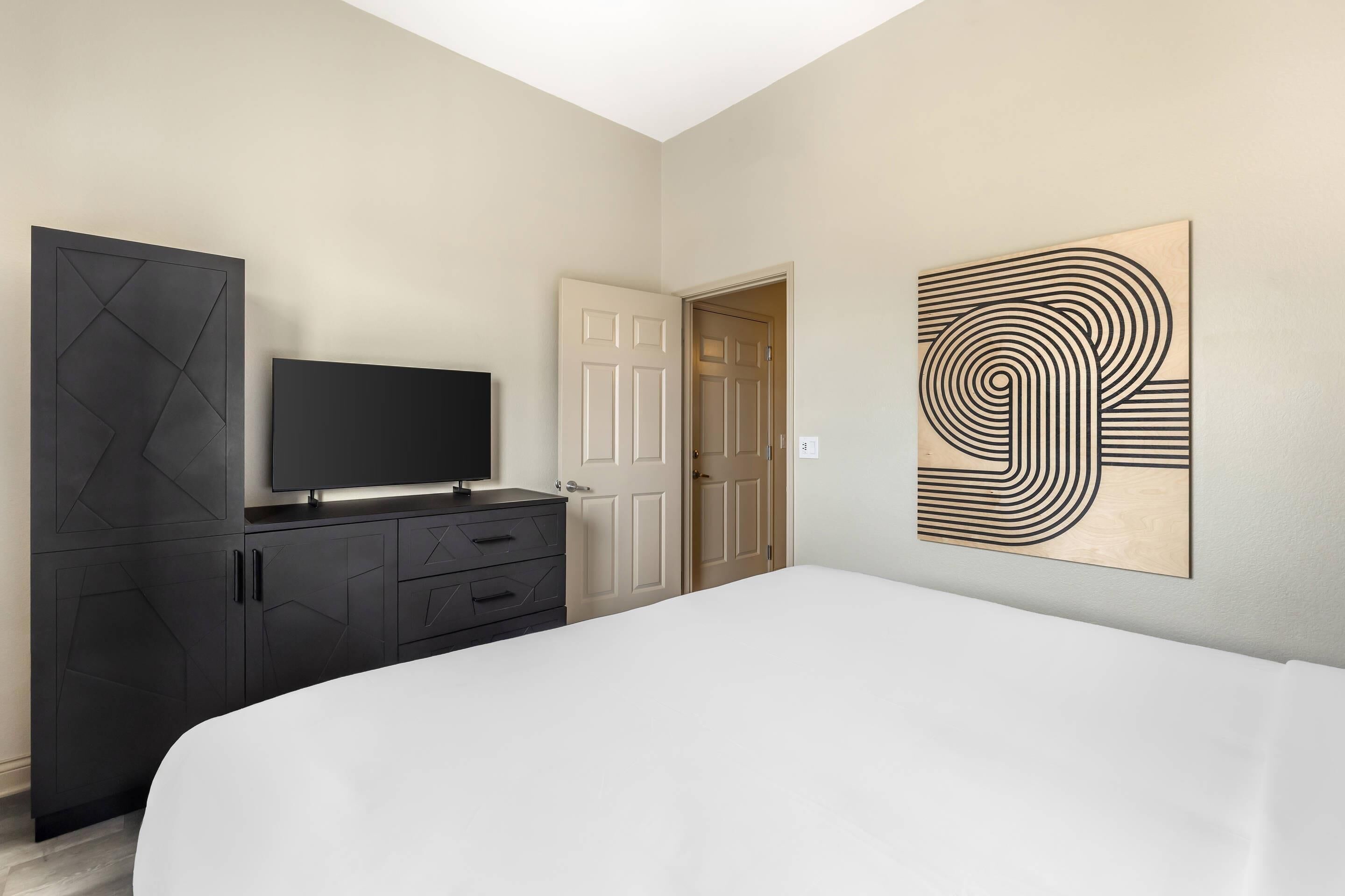 A bedroom with a bed, a TV on a dresser, a door, and a wall art piece.