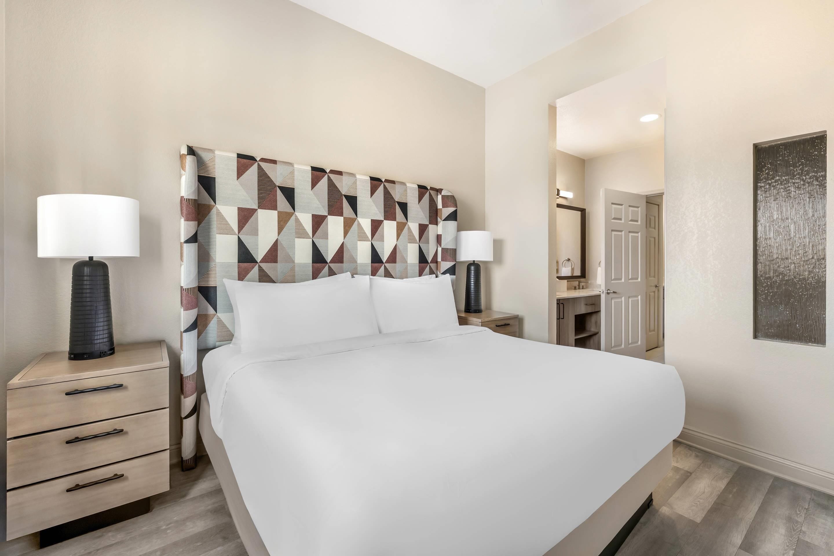 Ein Hotelzimmer mit einem großen Bett, zwei Nachttischen mit Lampen, einem Kopfteil mit geometrischem Muster, einem Spiegel und einer Tür zu einem weiteren Raum.