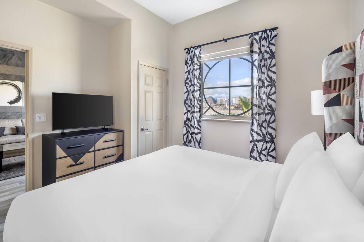 Habitación de hotel con cama, televisor, cómoda, ventana con cortinas estampadas y vista a la ciudad.