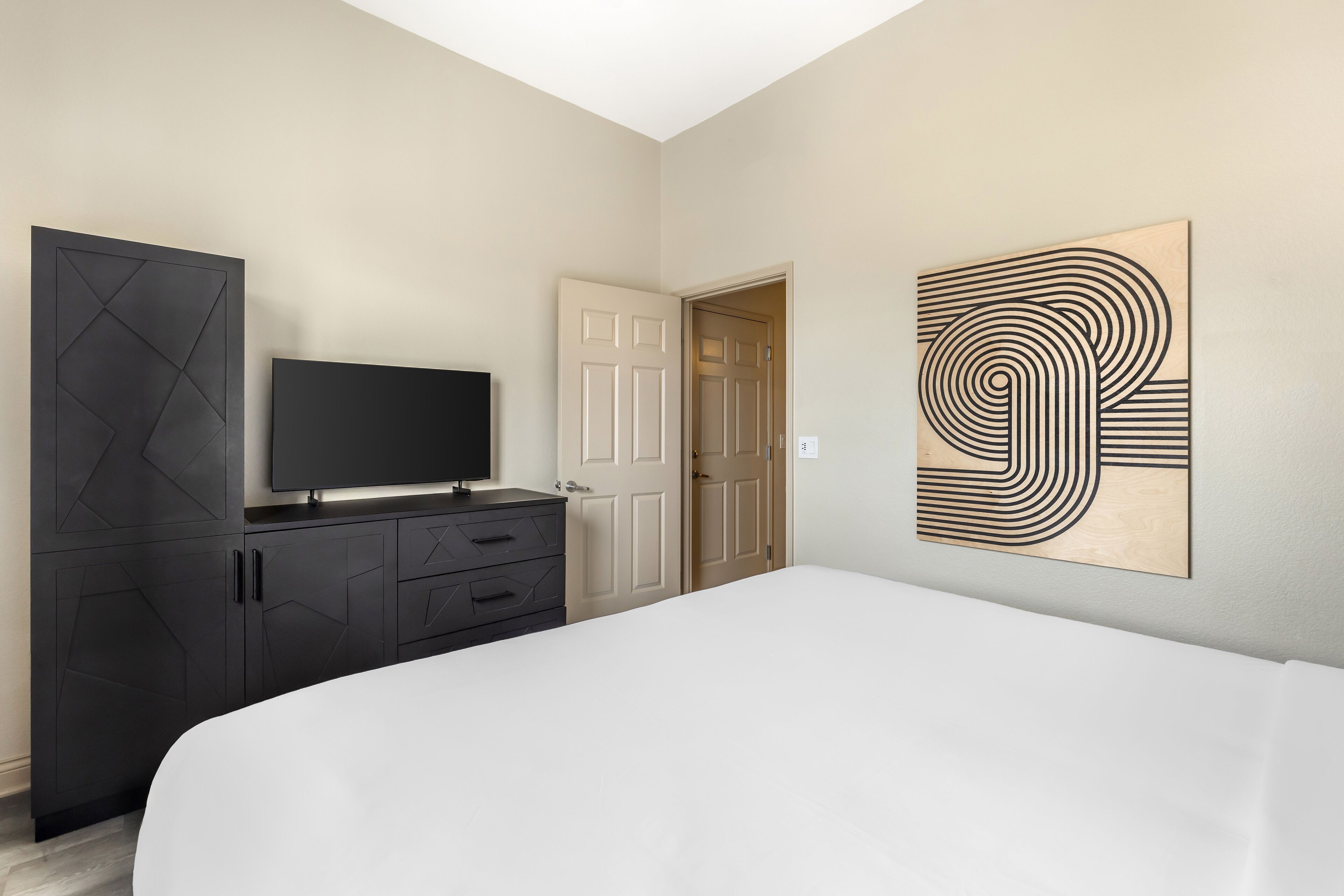 Un dormitorio con cama, televisor en una cómoda, una puerta y una obra de arte en la pared.