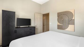 Un dormitorio con cama, televisor en una cómoda, una puerta y una obra de arte en la pared.