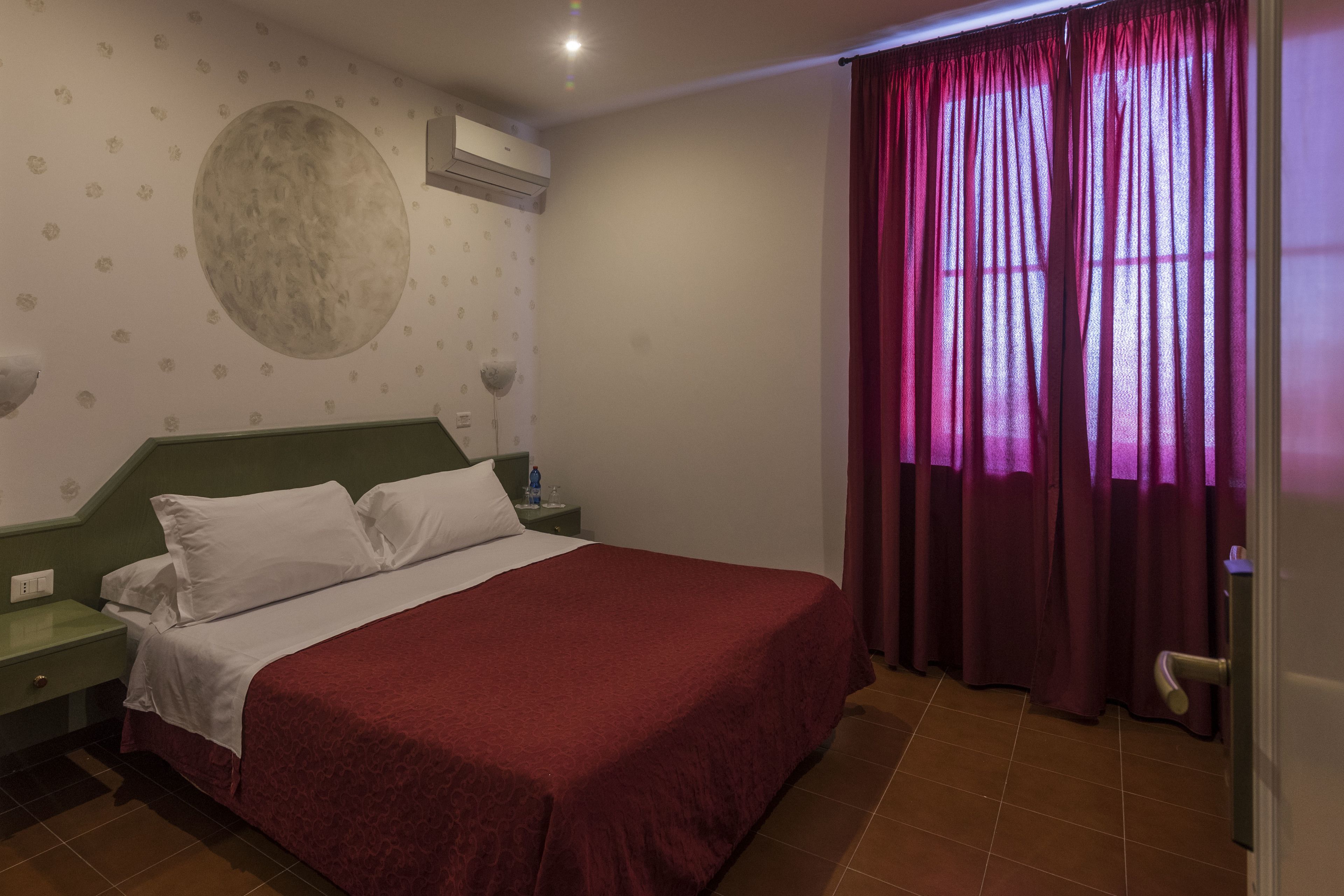 Ein Schlafzimmer mit einem Bett, einem grünen Nachttisch, weißen Kissen, einer roten Bettdecke, einer Wanduhr und einem Fenster mit violetten Vorhängen.