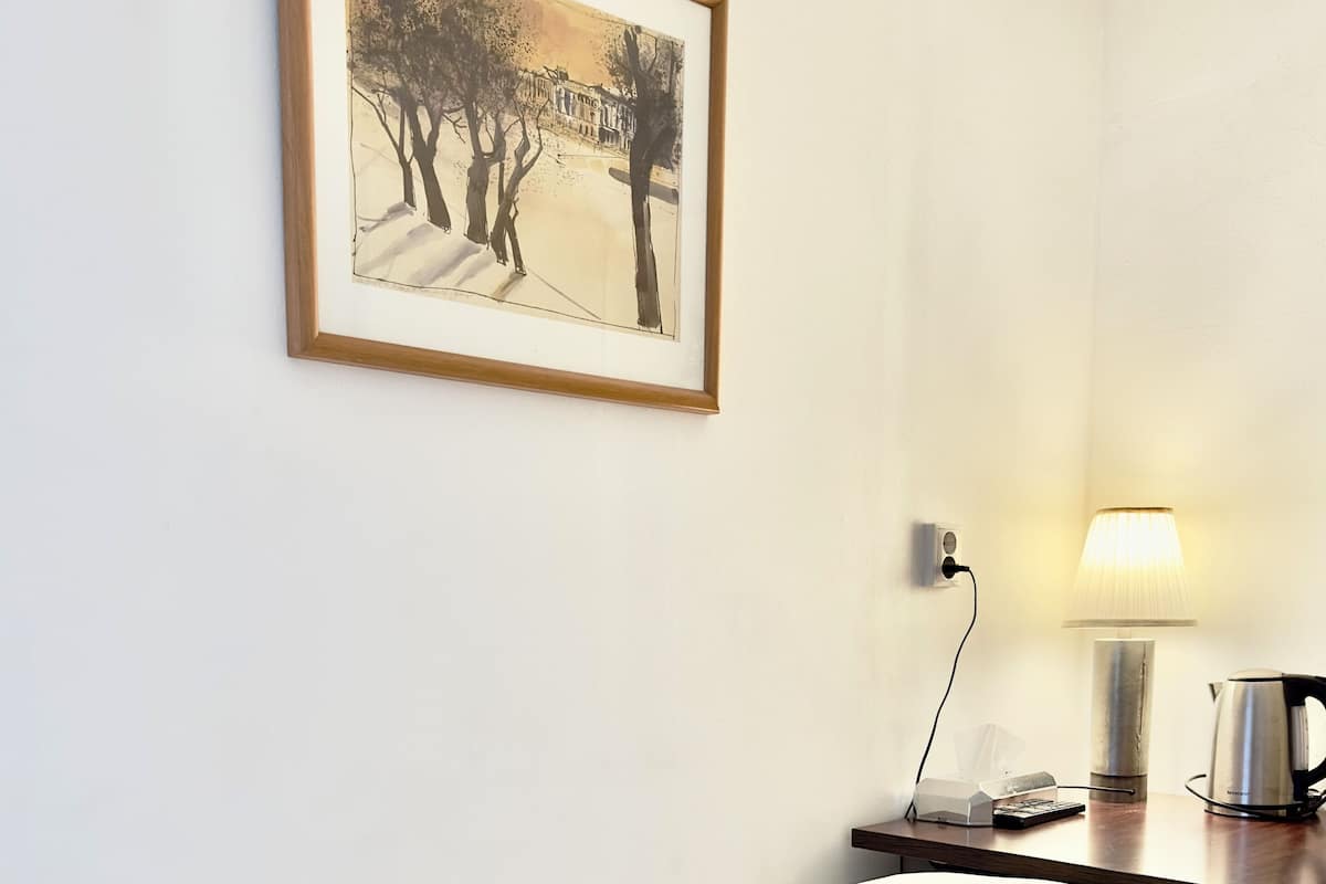 A hotel room with a bed, a bedside table with a lamp, a kettle, and a framed picture on the wall.