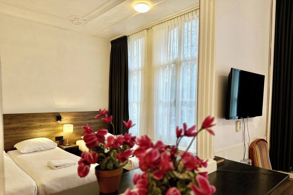 A hotel room with a bed, a bedside table with a lamp, a TV mounted on the wall, and a window with curtains.
