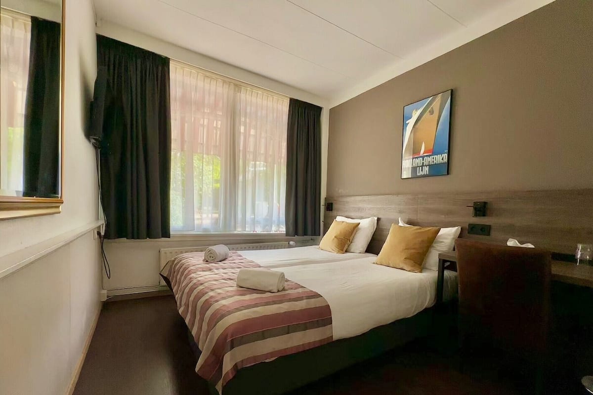 A hotel room with a bed, a window with curtains, a wall-mounted poster, and a chair.