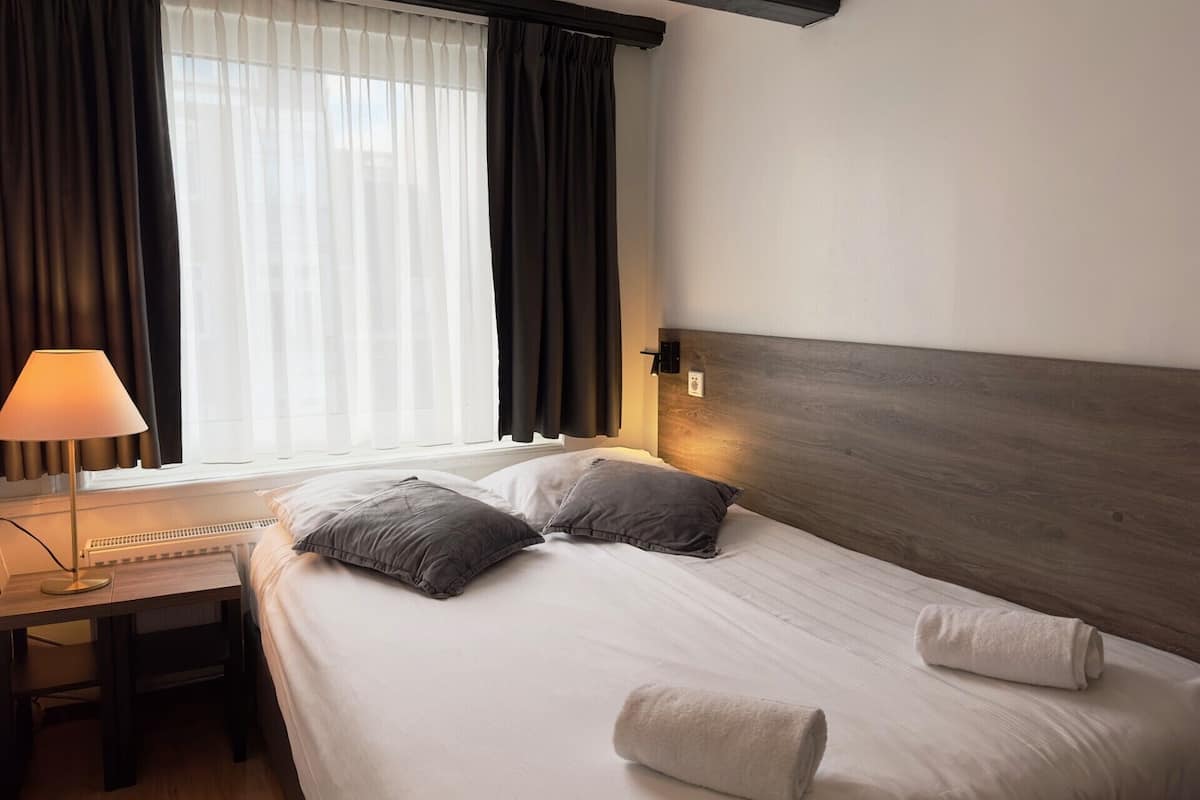 A hotel room with a bed, two pillows, a bedside lamp, a desk, and a window with curtains.