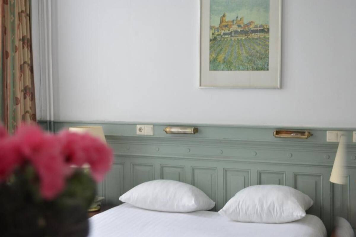 A hotel room with a bed, white pillows, a framed painting of a landscape, and a vase with pink flowers.