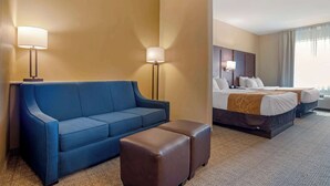 A hotel room with a blue sofa, a brown ottoman, a bed with two pillows, and a nightstand. - Comfort Suites Alexandria North (Alexandria)