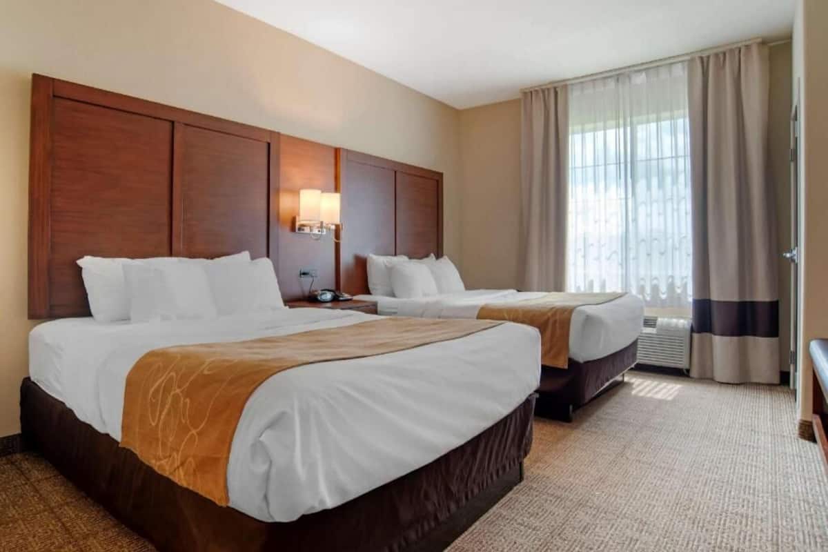 Une chambre d’hôtel avec deux lits, une grande tête de lit en bois, une table de chevet avec une lampe et une fenêtre avec des rideaux.