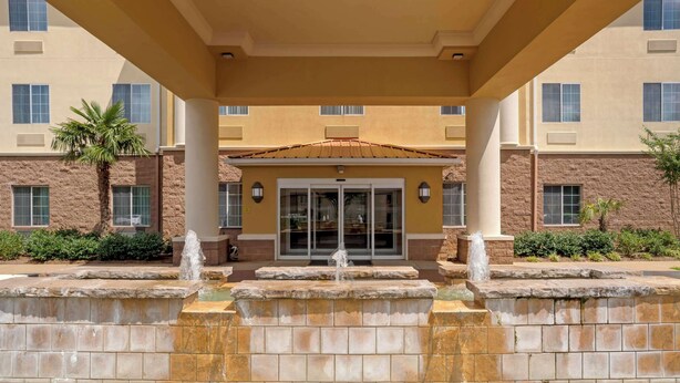 A hotel entrance with a stone fountain, a covered walkway, and a building with multiple windows and a palm tree. - Comfort Suites Alexandria North (Alexandria)