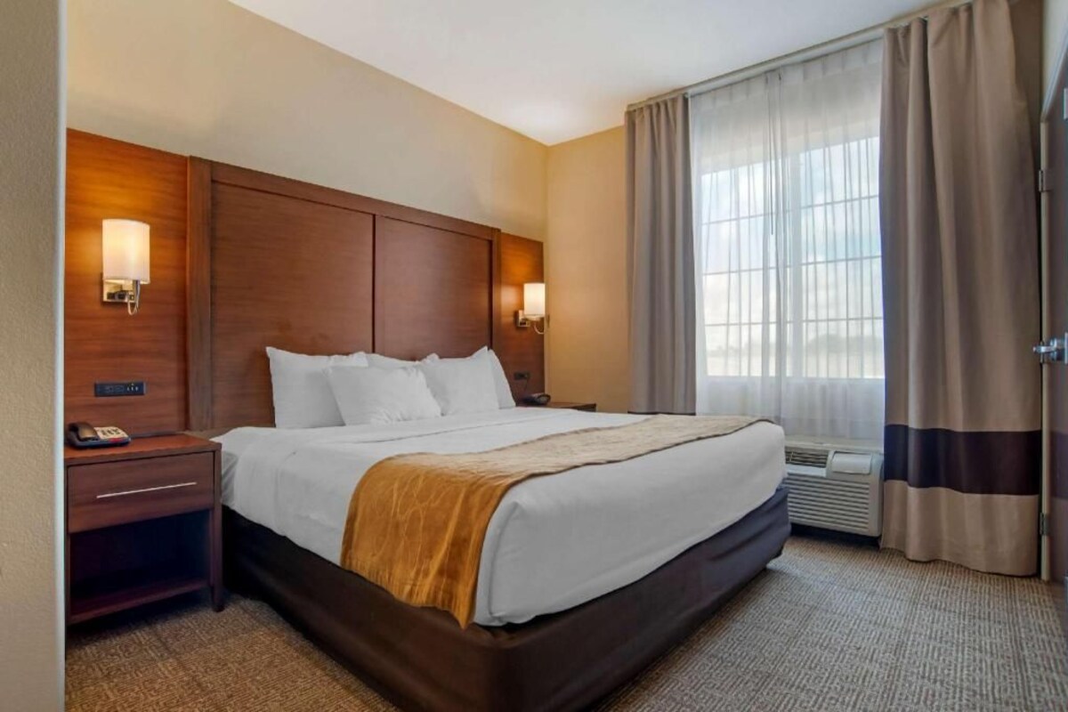 A hotel room with a large bed, bedside lamps, a nightstand, a window with curtains, and an air conditioning unit.