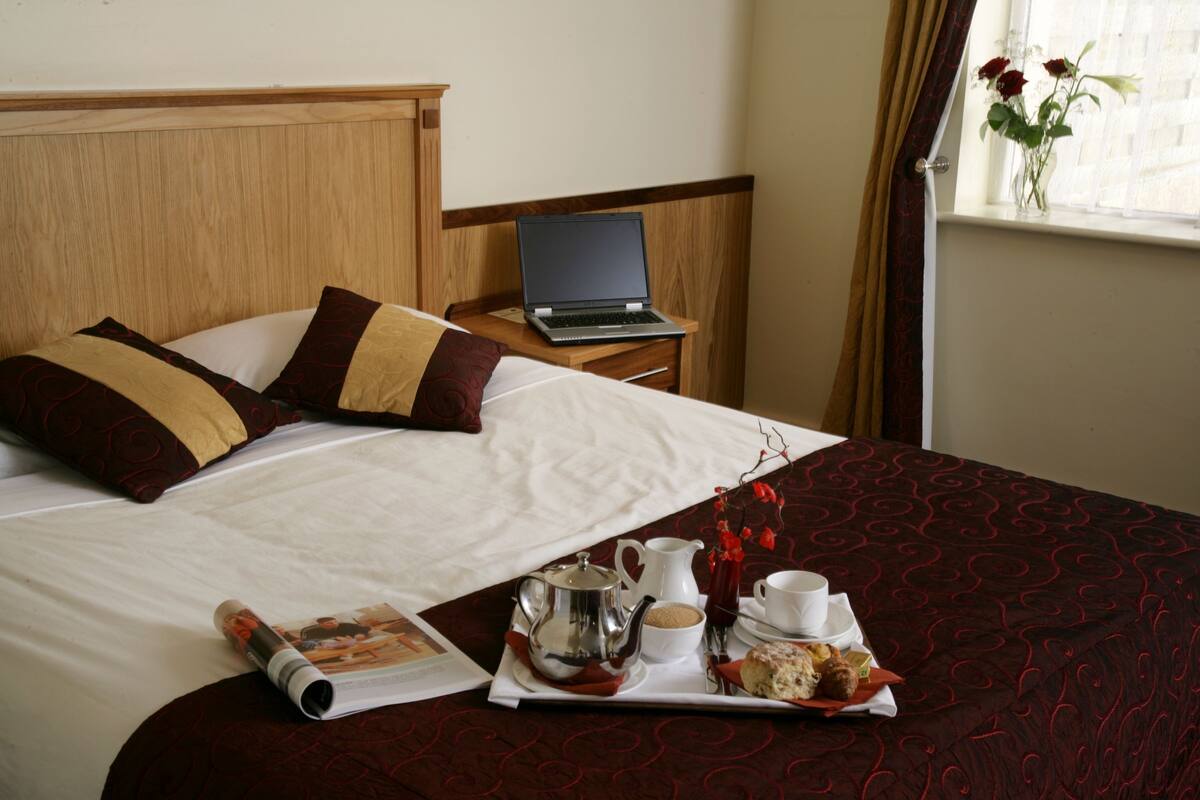 A hotel room with a bed, a laptop on a bedside table, a tray with breakfast items, and a window with flowers.
