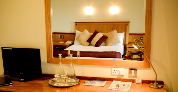 Habitación de hotel con cabecera de madera, cama con almohadas, mesita de noche con lámpara, bandeja con botellas y una revista.