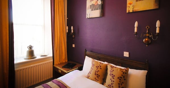 A hotel room with a bed, a window with curtains, a radiator, a bedside table with a book, and wall-mounted lamps. - Bush Hotel (Carrick-on-Shannon)
