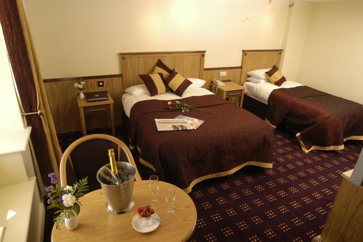 A hotel room with two beds, a small table with a bottle of champagne, a vase with flowers, and a newspaper.