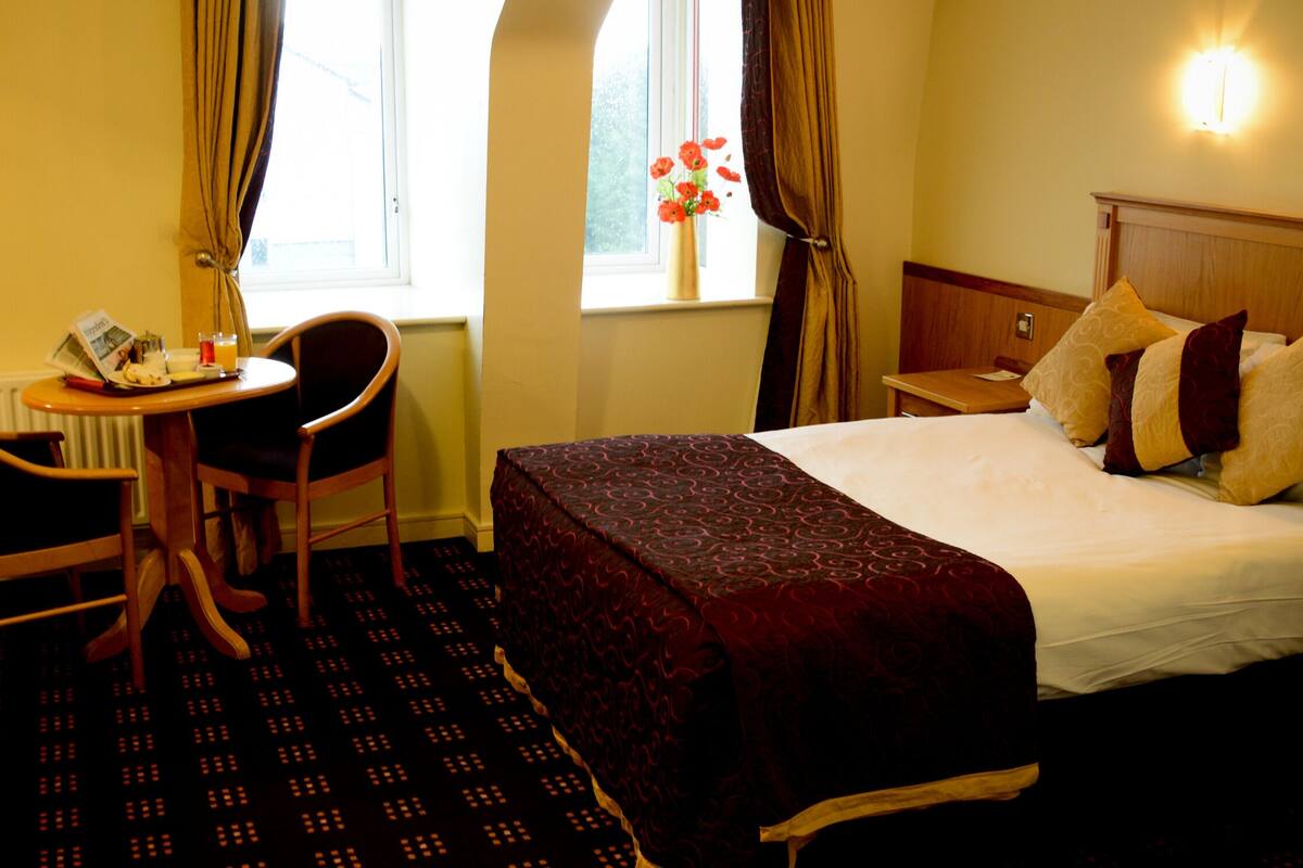 A hotel room with a bed, a desk with a chair, a small table, and a window with curtains.