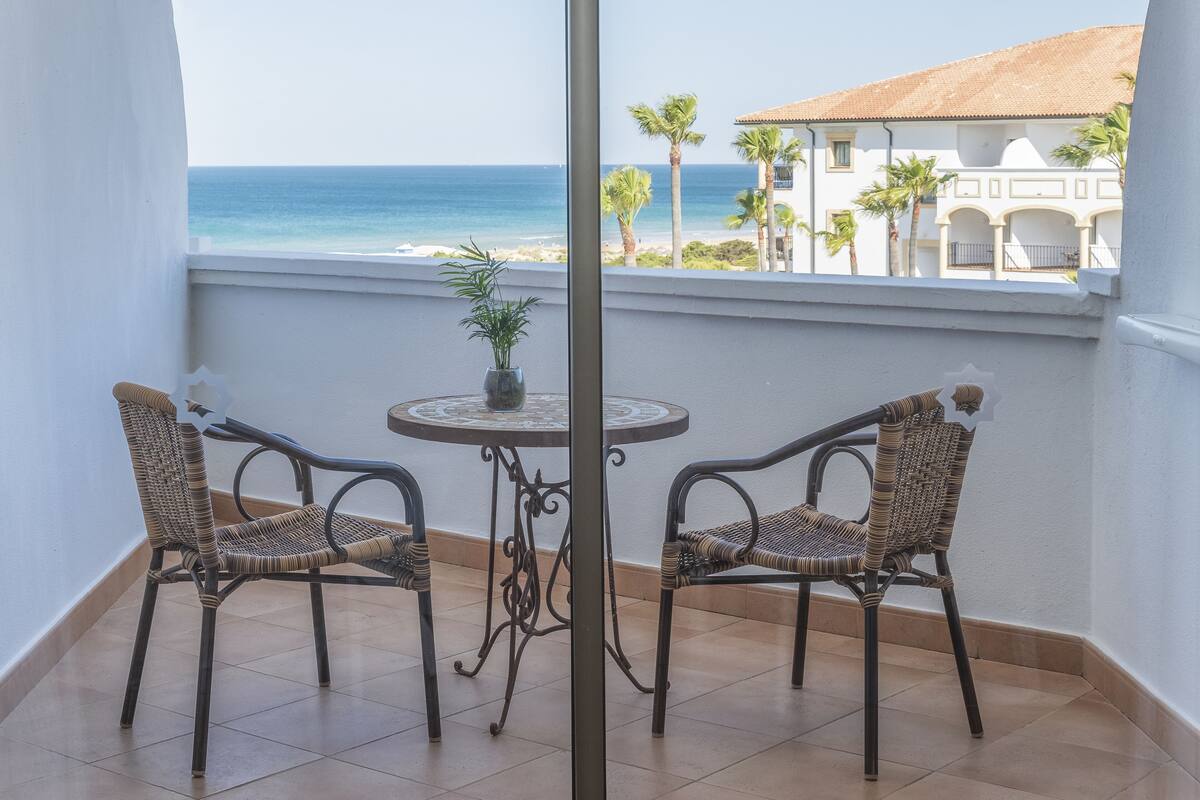 Un balcón con dos sillas de mimbre y una mesita pequeña, con vista a una playa y palmeras.
