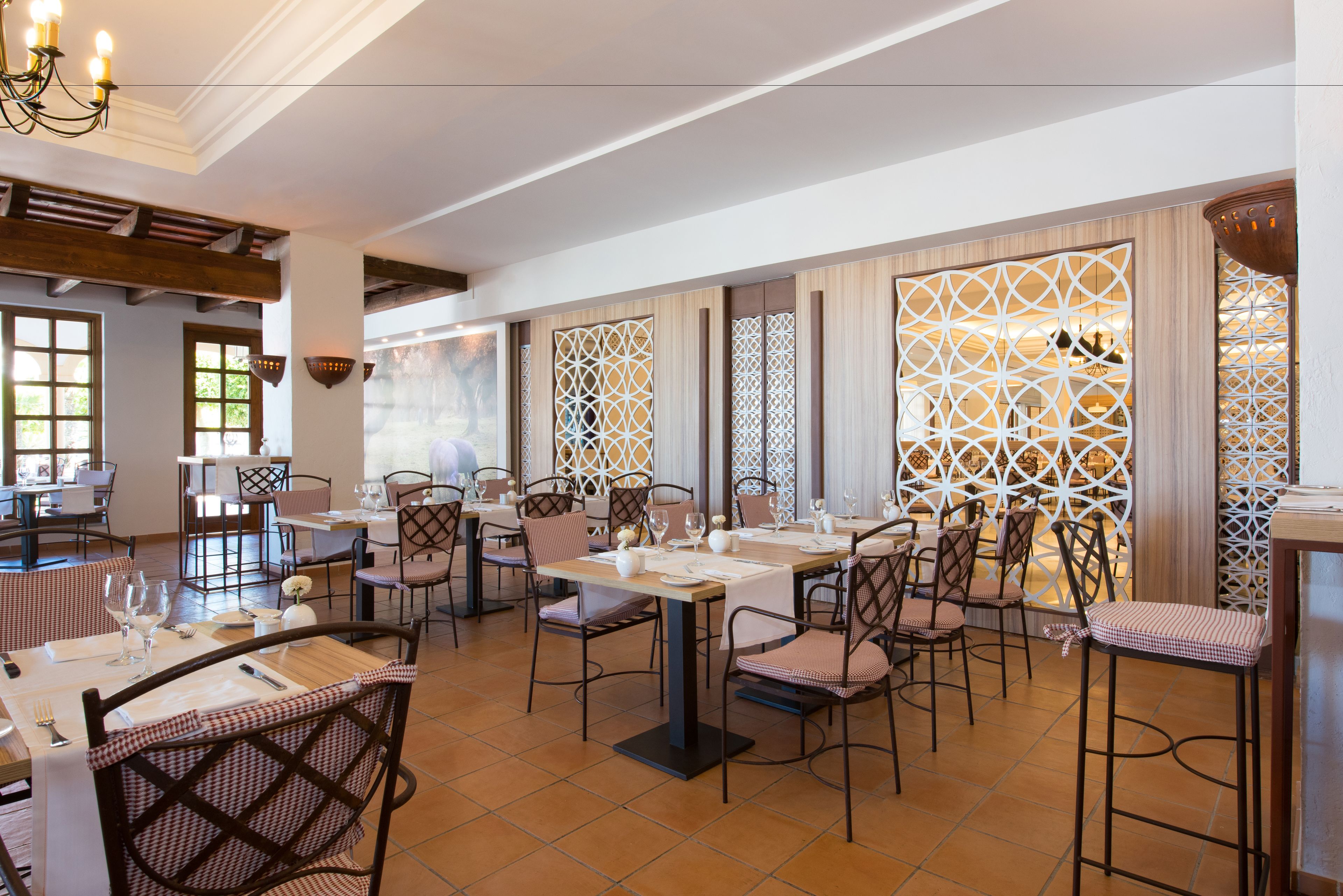 Um restaurante com várias mesas e cadeiras, vigas de madeira no teto e painéis de treliça decorativa nas janelas.