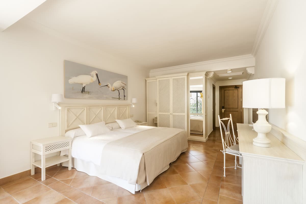 Un dormitorio con cama, mesita de noche, escritorio con lámpara, silla y puerta que da a otra habitación.