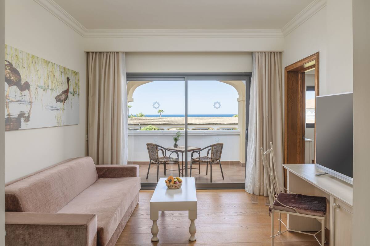 Una habitación con sofá, una mesa con fruta, televisión y un balcón con sillas y vistas al mar.