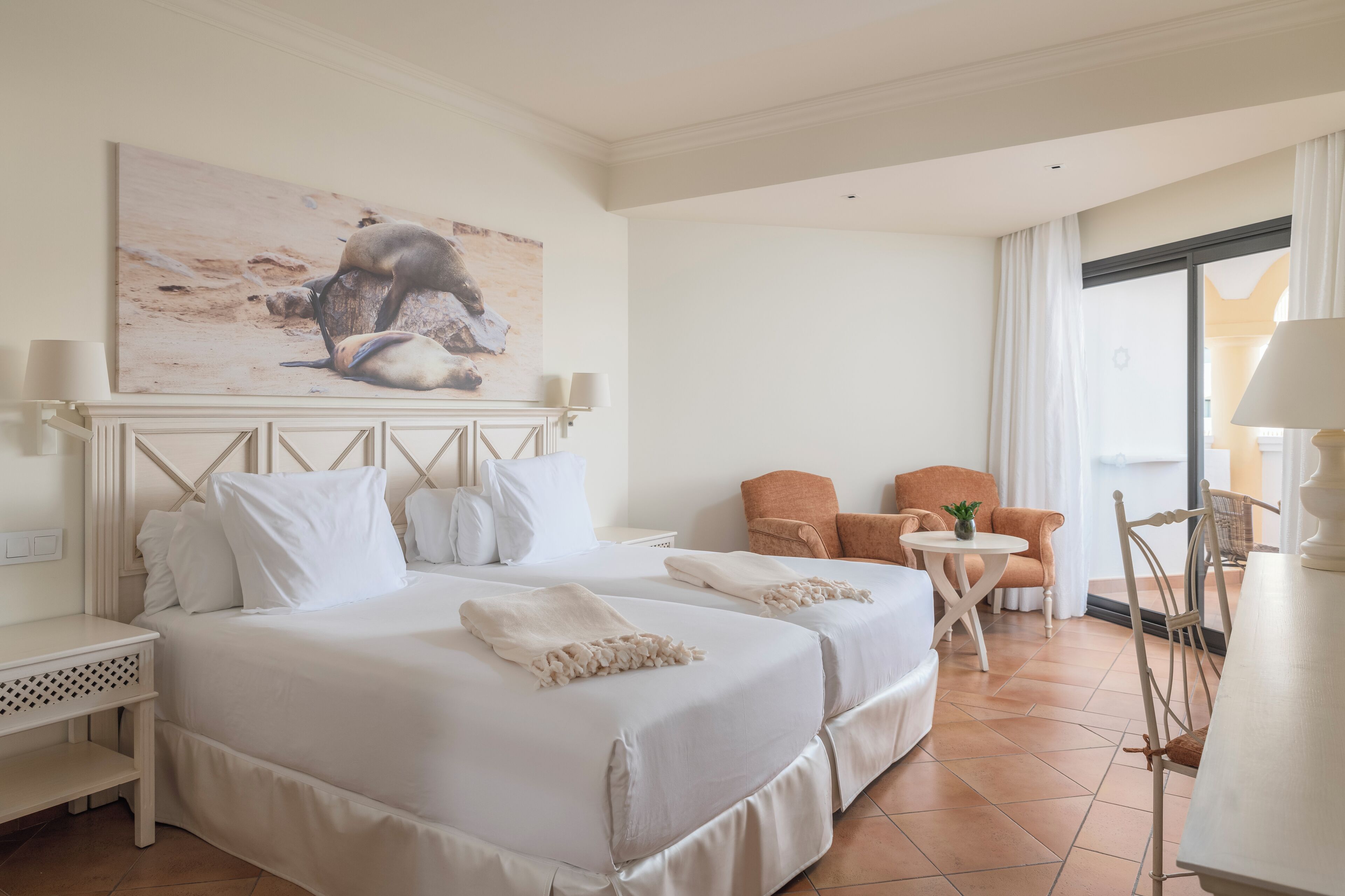 Una habitación de hotel con dos camas, un cuadro, una silla, una mesa y una lámpara.