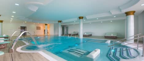 Una piscina interior con agua cristalina, rodeada de suelo de madera y paredes blancas.