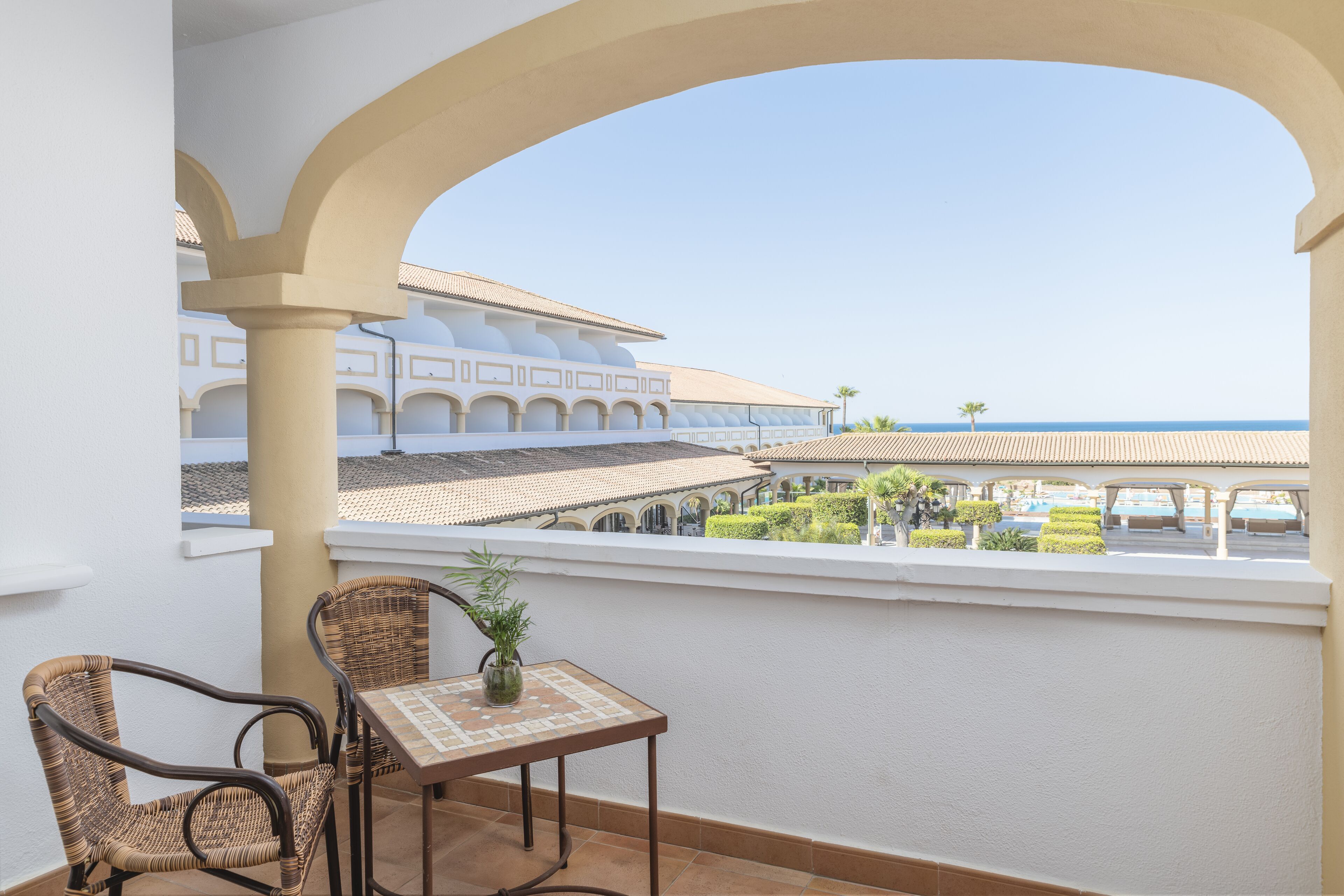Een balkon met rieten stoelen, een kleine tafel en een bloempot. Het balkon heeft uitzicht op een zwembad met een wit gebouw op de achtergrond.