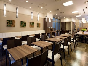 A modern restaurant interior with long wooden tables and black chairs, recessed ceiling lighting, and wall-mounted artwork.