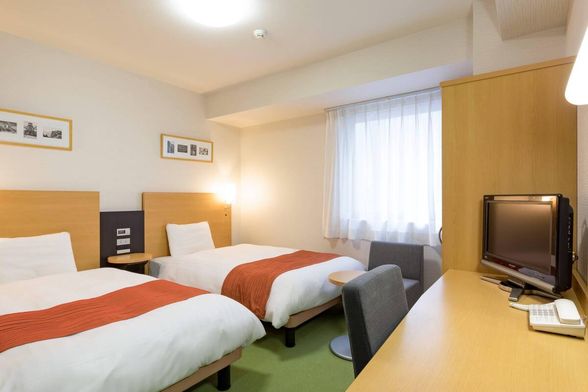 A hotel room with two beds, a desk with a computer and phone, a chair, and a window with curtains.